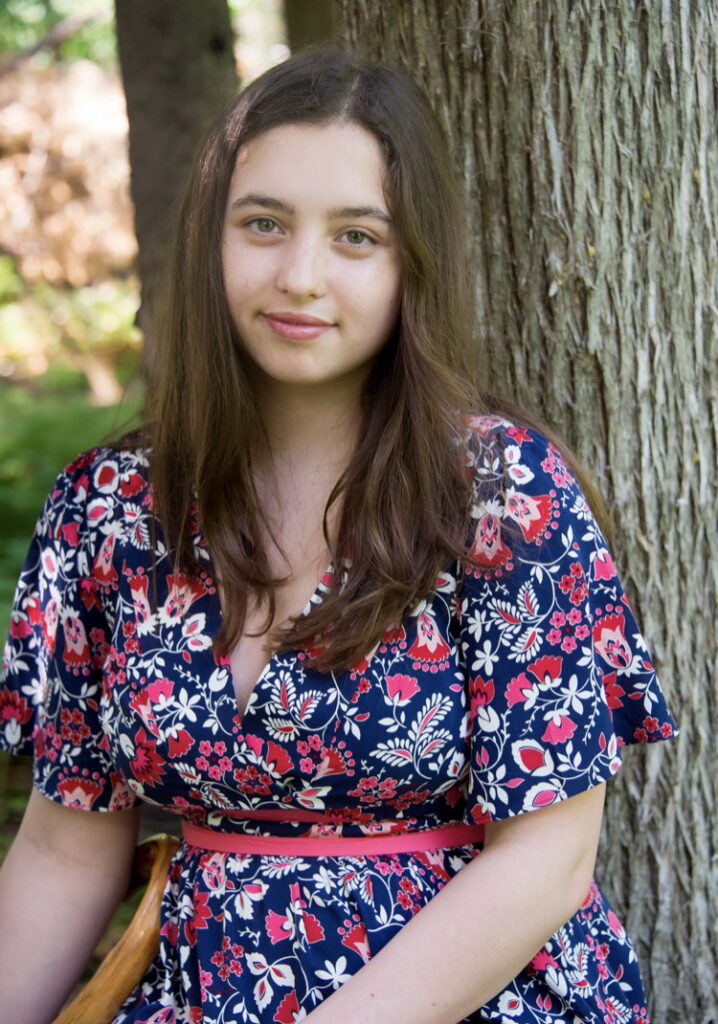 Image Description: Izzy is wearing a blue dress with pink and white flowers and leaves.  She is looking straight at the camera with a slight smile and sitting in front of some trees.