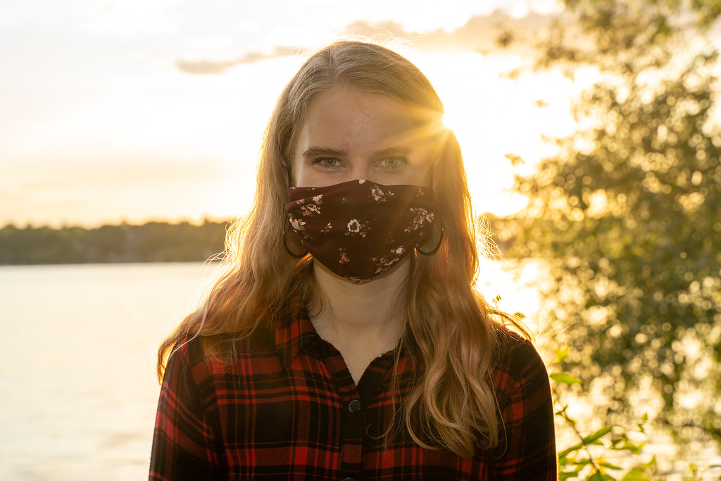 Image Description: Eleanor is wearing a red and black plaid flannel shirt and a burgundy mask with light pink flowers on it.  She is looking straight at the camera and standing in front of a lake.