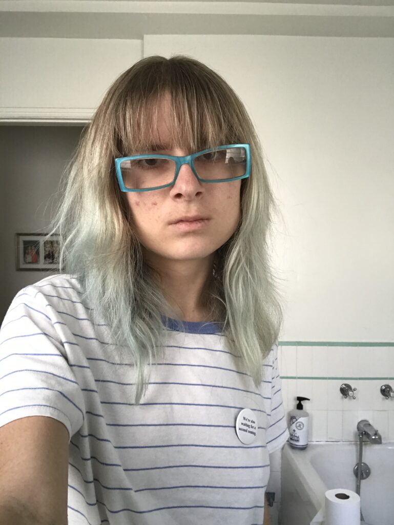 Image Description: Z wearing a white t-shirt with blue horizontal stripes.  She is holding the camera with her hand and looking straight at the camera with a straight face.