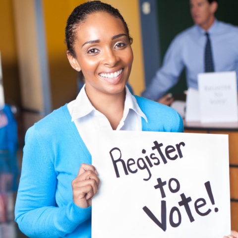A Black woman wearing a white collared button-up and a light blue cardigan is smiling and holding a sign that reads: Register to Vote1